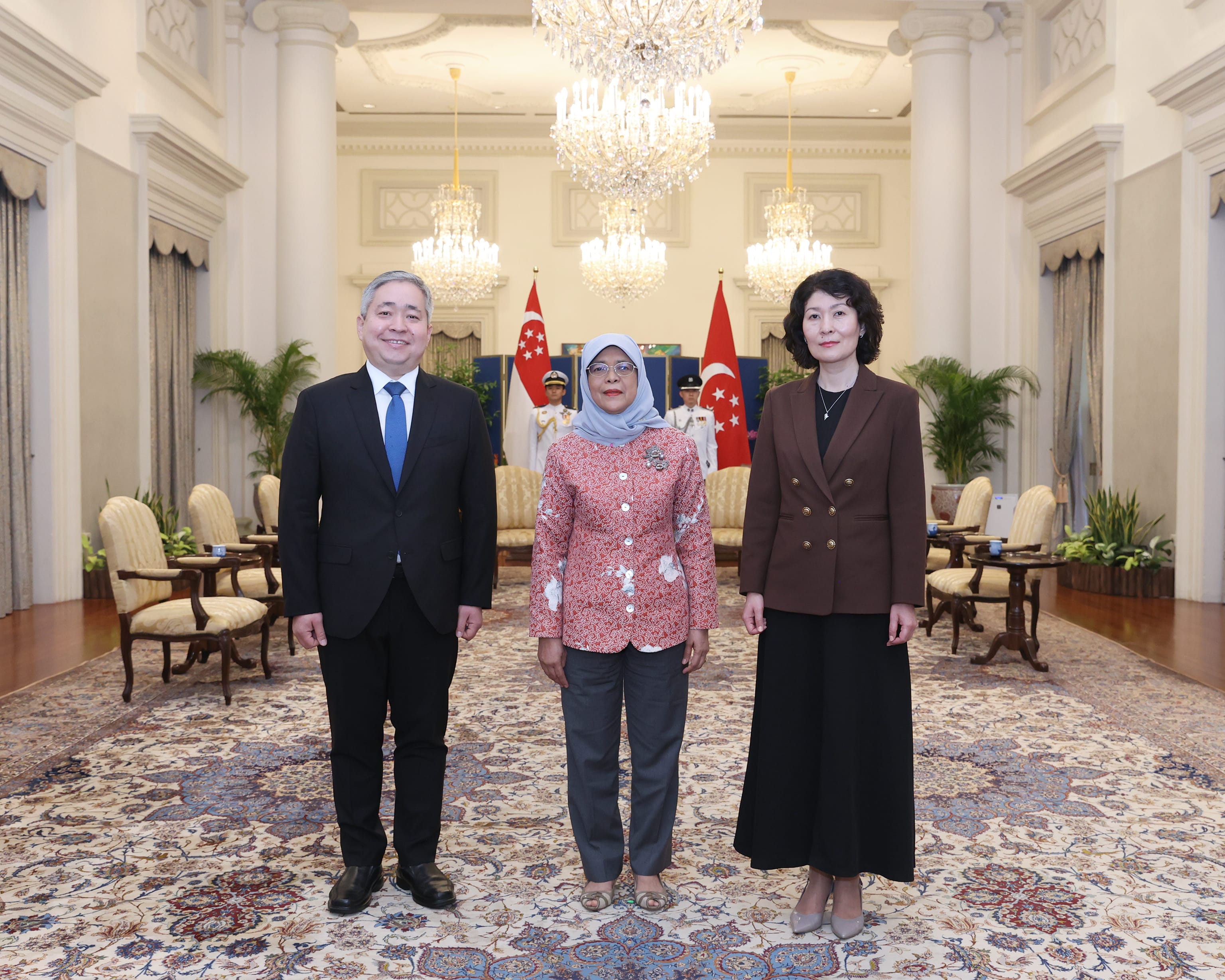 Three people stand indoors, with Singapore flags behind. Halimah Yacob is in the center.